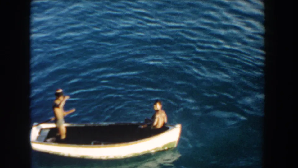 1960 Miami home-movie frame of a small white boat moving across rippled blue water, with one person standing and another seated.