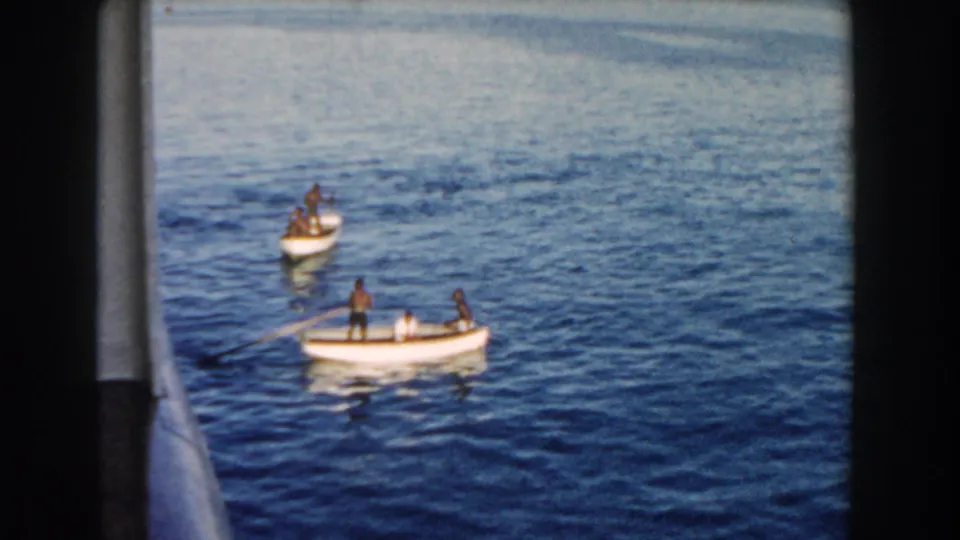 1960 Miami frame of two small rowboats separated on calm blue water, with distant boaters visible from a vessel window.