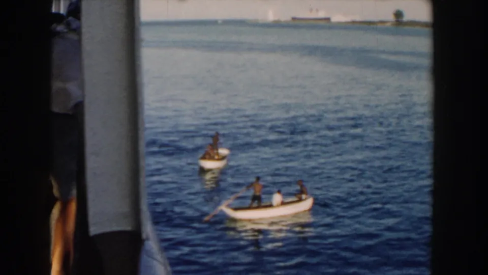 1960 Miami home-movie frame showing a rowboat with several people and a visible oar, with another small boat nearby and shoreline in the distance.