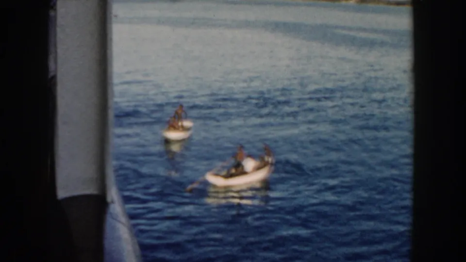 1960 Miami frame showing two small rowboats with people on calm water, seen through the edge of a boat window with faded color and grain.