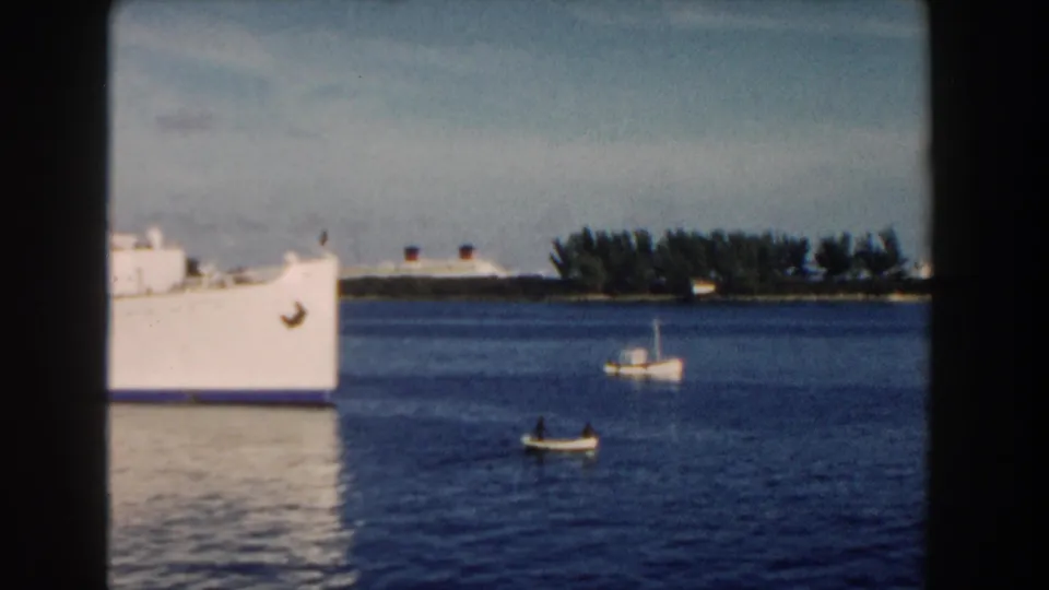 The bow of a white passenger ship fills the left side while two small boats sit farther out in 1960 Miami harbor footage.