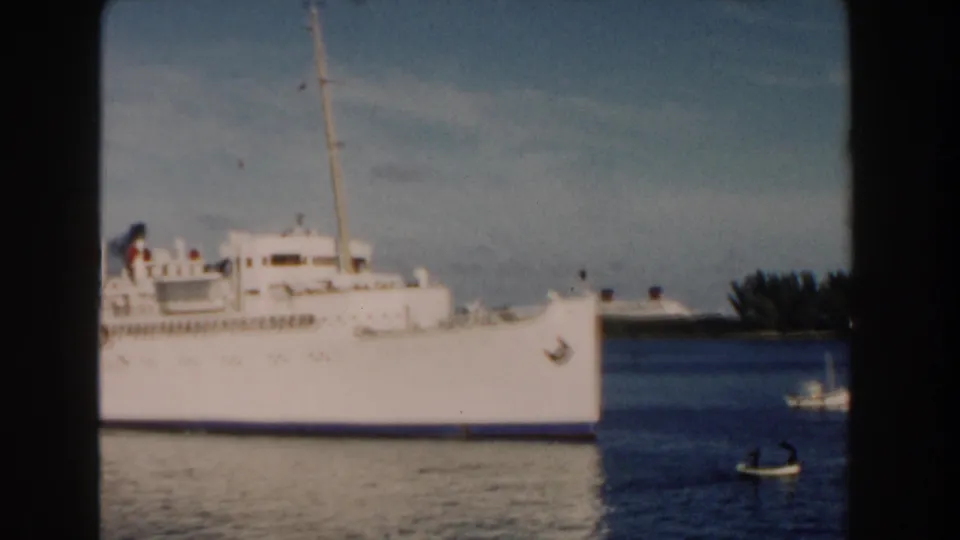 1960 Miami harbor view of a large white passenger ship moving past calm water, with a small boat and distant shoreline visible.