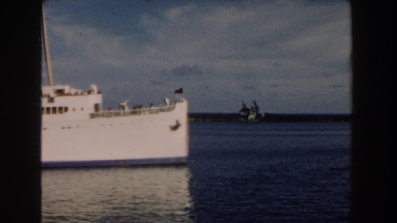 1960 Miami 8mm frame showing the bow and forward side of a large white passenger ship on calm water, with a distant shoreline behind it.