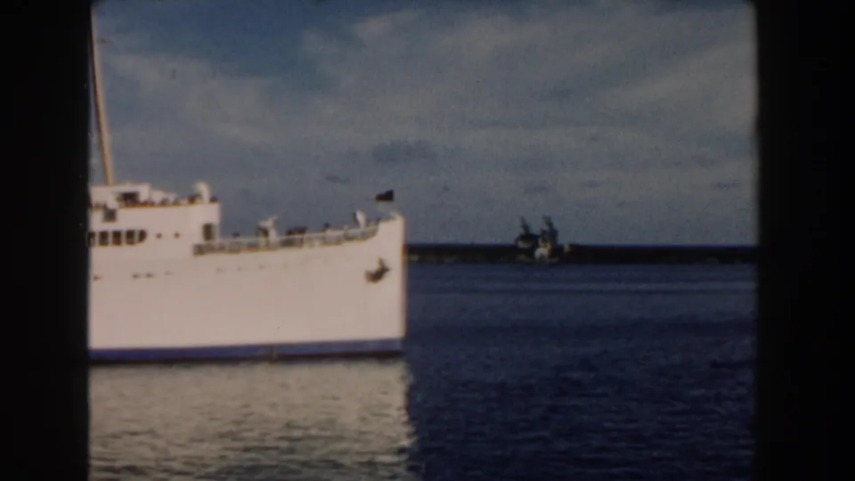 1960 Miami 8mm frame showing the bow and forward side of a large white passenger ship on calm water, with a distant shoreline behind it.