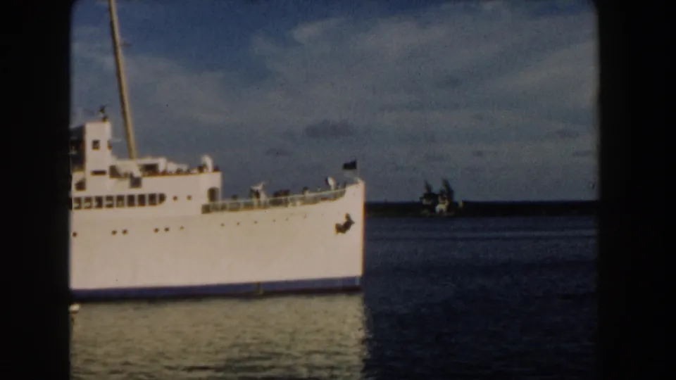 1960 Miami home-movie frame of a white passenger ship passing broadside, with deck railings, water reflections, and a small distant boat visible.