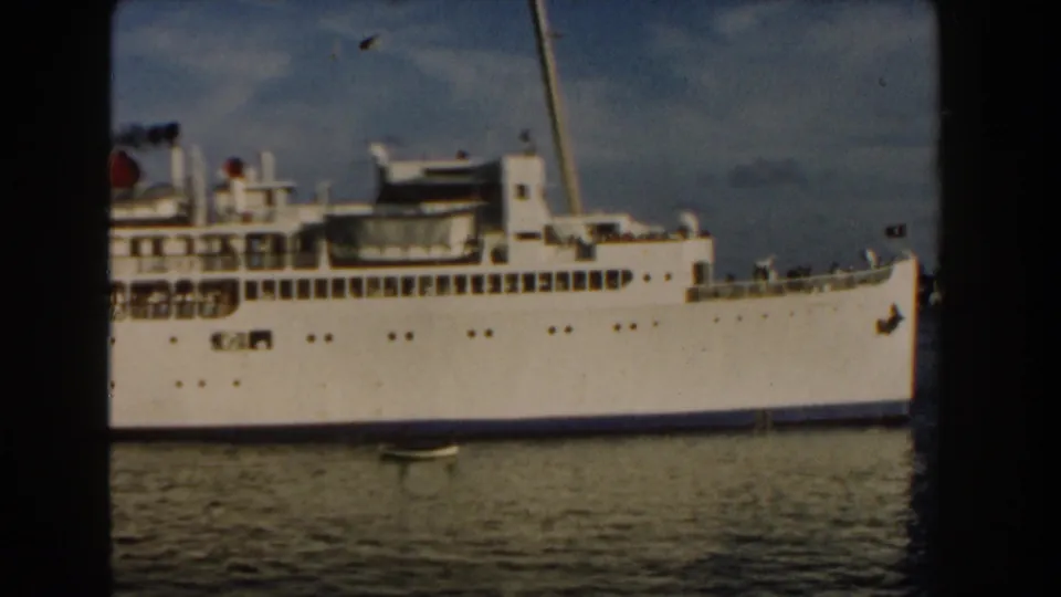 1960 Miami frame of a large white passenger ship filling the view, with visible deck structures, railings, tiny passengers, and calm water in front.