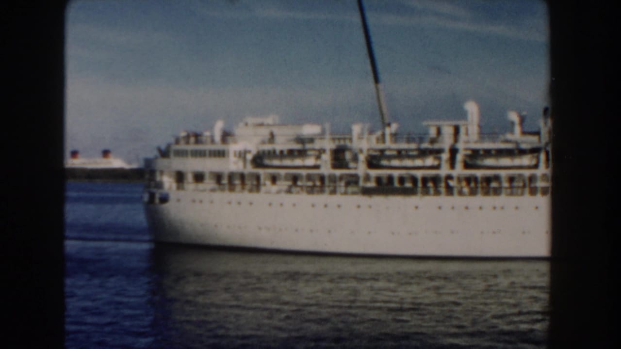 Side of a white cruise ship with decks and funnel, seen through dark vertical film borders.