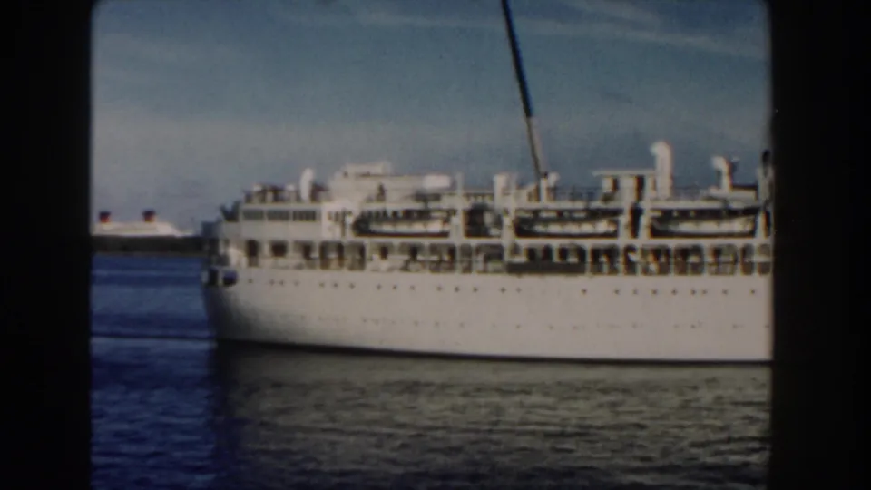 1960 Miami-area 8mm view of a white cruise ship sliding past, with upper decks, lifeboat shapes, funnel markings, and blue water visible.