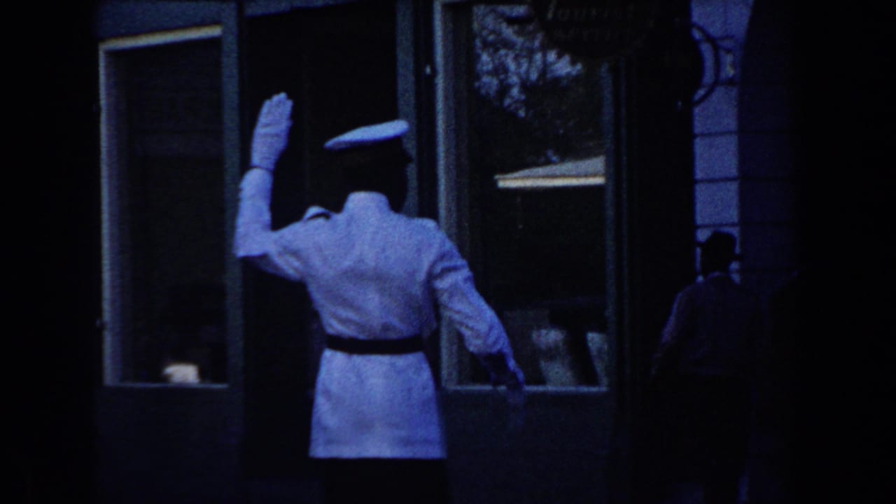1960 Miami 8mm frame showing a white-uniformed traffic officer from behind as he signals near a commercial building entrance.