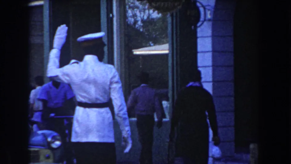 1960 Miami street view with a traffic officer in white directing movement while pedestrians pass near storefront windows.