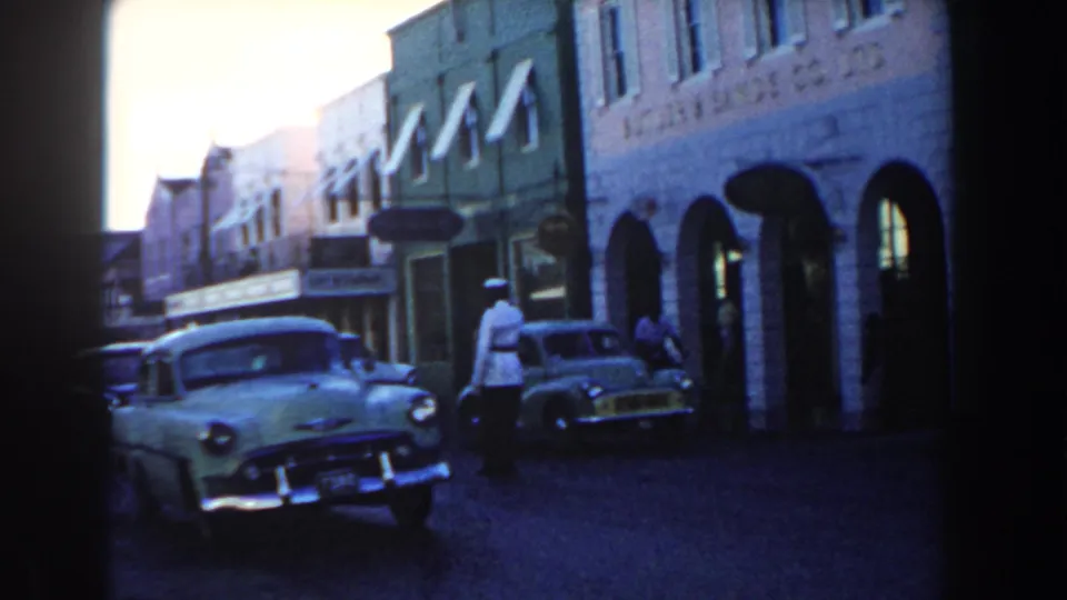 1960 Miami 8mm street scene with mid-century cars, brick storefronts, awnings, and a traffic officer near the curb.
