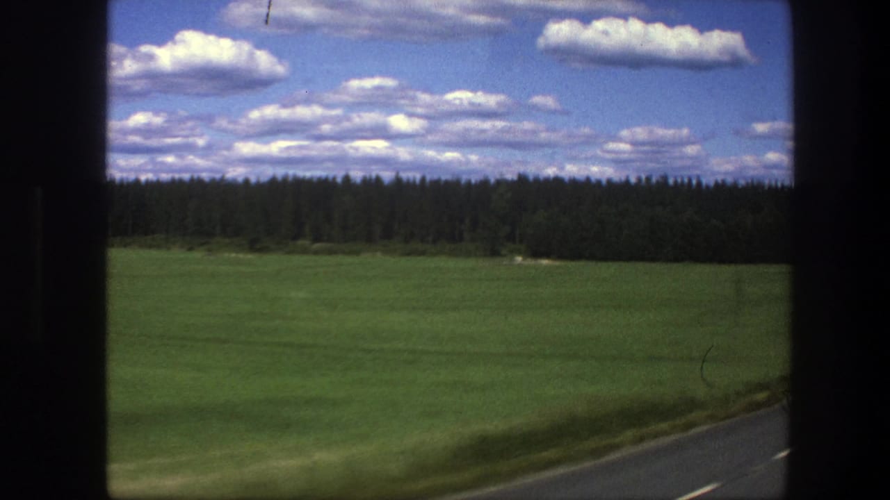 1975 Swedish countryside view from a moving vehicle, showing a grassy field, roadside, forest edge, and scattered clouds.
