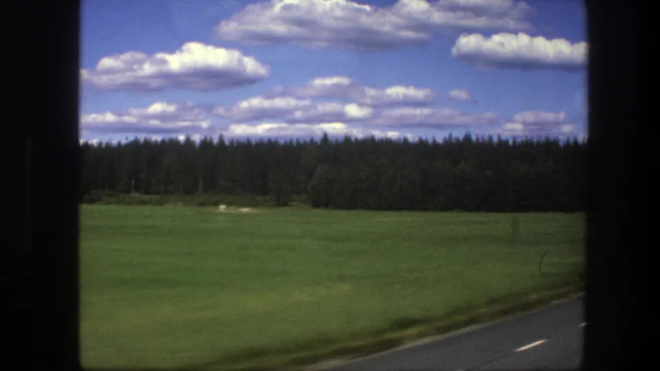 Rural Swedish road view from 1975 home-movie footage, showing field, forest, road markings, and a bright cloud-filled sky.