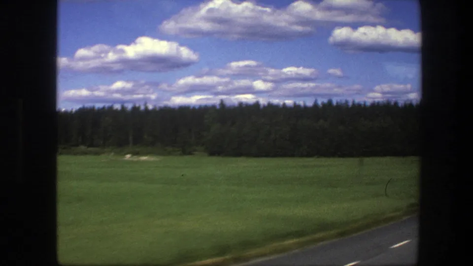 1975 Swedish home-movie frame showing a green field, distant forest, paved roadside, and bright clouds seen from a moving car.