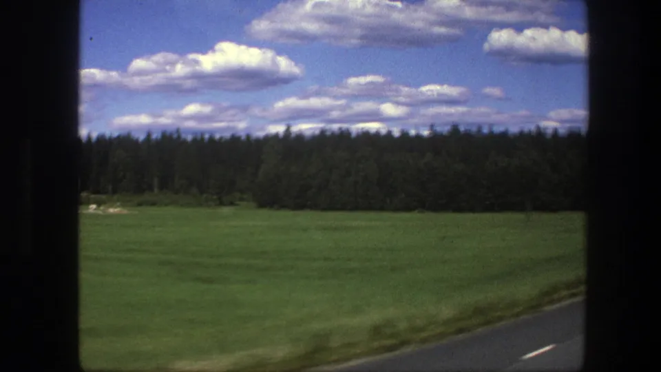 Moving car-window view in Sweden, 1975, with a broad green field, dense forest horizon, road shoulder, and scattered clouds.