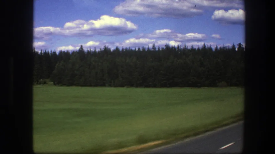 Swedish 1975 home-movie view from a moving car, with roadside pavement, green field, forest, and bright clouds in frame.