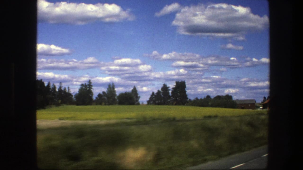 1975 Swedish rural landscape from a vehicle window, showing fields, trees, roadside vegetation, and small distant buildings.