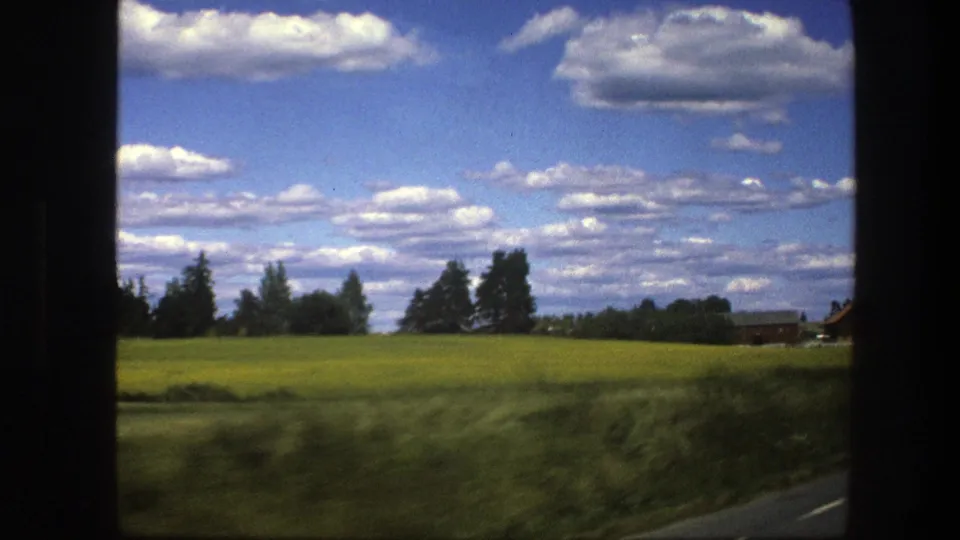 1975 Swedish countryside frame with a yellow-green field, roadside blur, trees, and a small distant rural building.