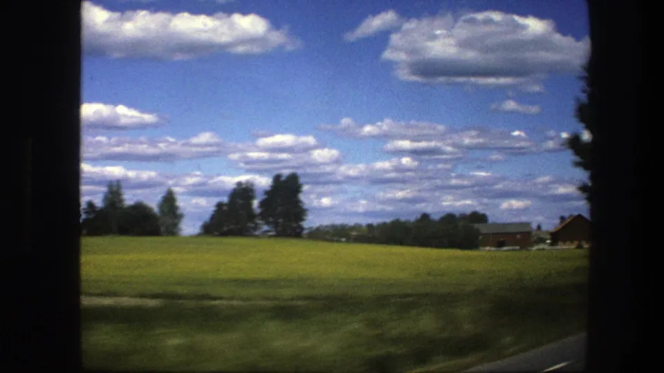 1975 Swedish road-view frame with open fields, distant trees and buildings, a road edge, and a bright sky with many clouds.
