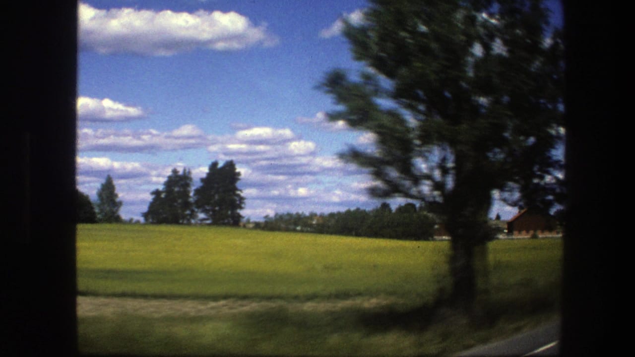 Green field and red farm buildings seen from a moving rural road.