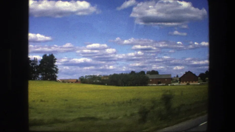 1975 Sweden countryside frame with green fields, low red farm structures, tree line, and scattered clouds viewed from the road.