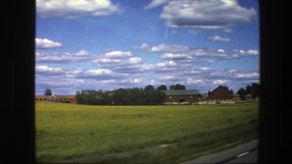 1975 Sweden 8mm frame showing a green field, red farmhouse or farm structures, trees, and the edge of a rural road.