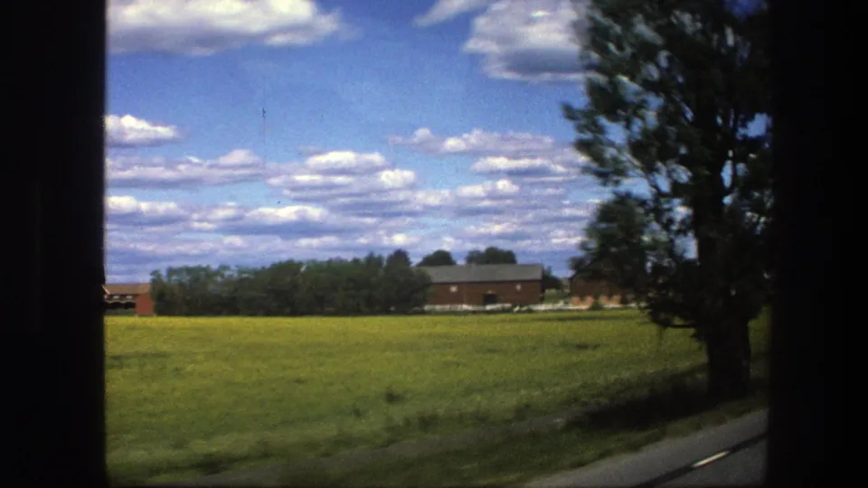 1975 Sweden rural road view with a passing tree, green field, red farm buildings, and a bright sky with scattered clouds.