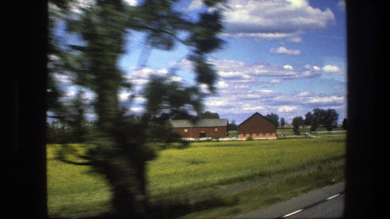 Green fields and red farm buildings seen from a moving vehicle, with trees blurred at the left.