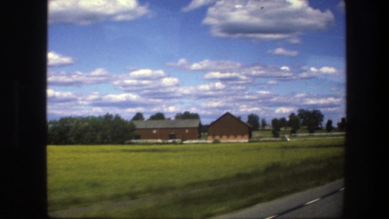 Red farm buildings across green fields under a blue sky with scattered white clouds.