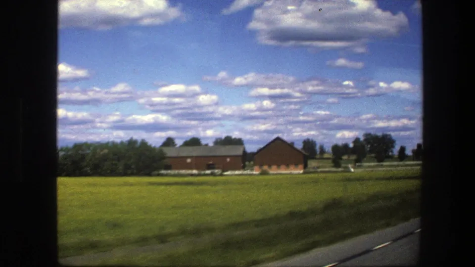 Rural Swedish fields and red farm structures appear beyond the roadside, framed by the dark edge of a vehicle window.