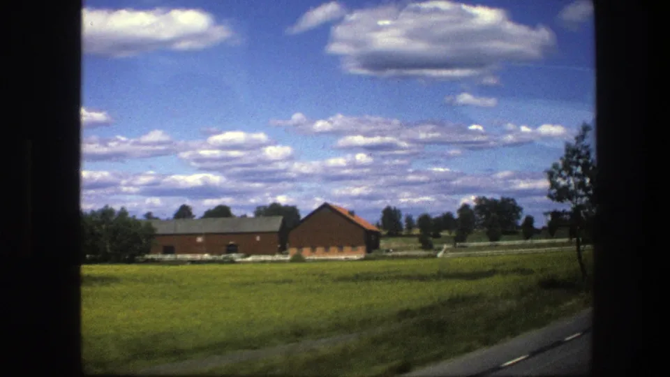 A passenger-window view shows Swedish countryside fields, a red farm building, roadside pavement, trees, and broad summer clouds.