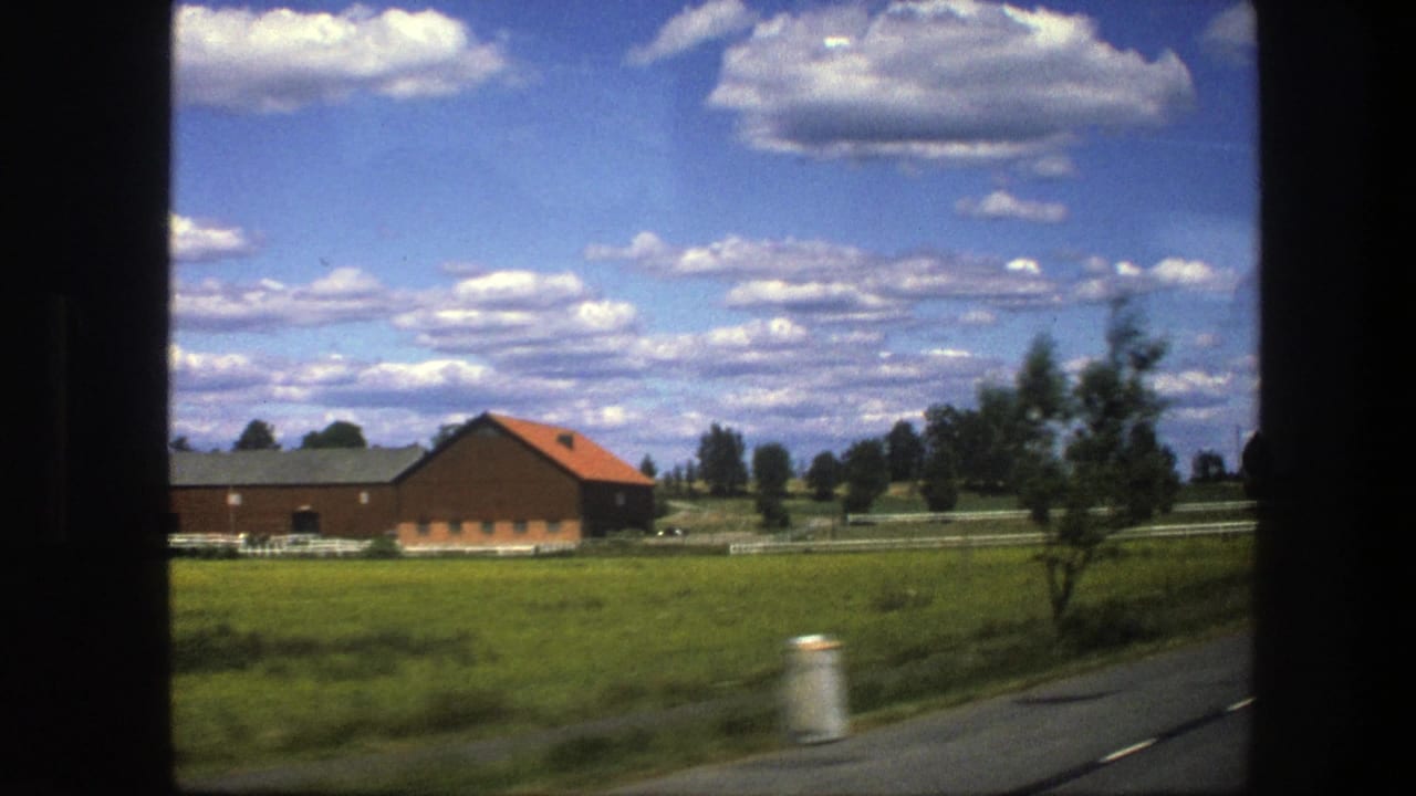 1975 Swedish countryside seen from a moving train, with a red-roofed barn, fenced field, roadside, and scattered clouds.