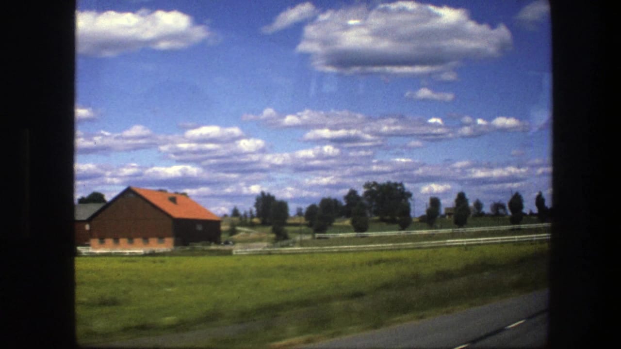 A Swedish rural scene from the train window shows a barn, pale fence lines, green fields, and a bright clouded sky.