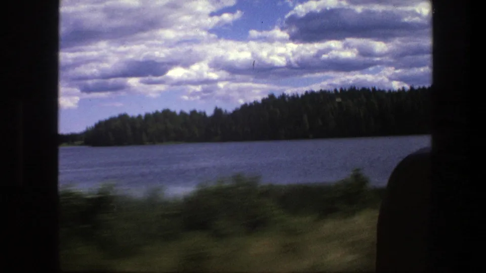 A lake and evergreen shoreline appear from the moving train, with blurred foreground vegetation and a cloudy sky above.
