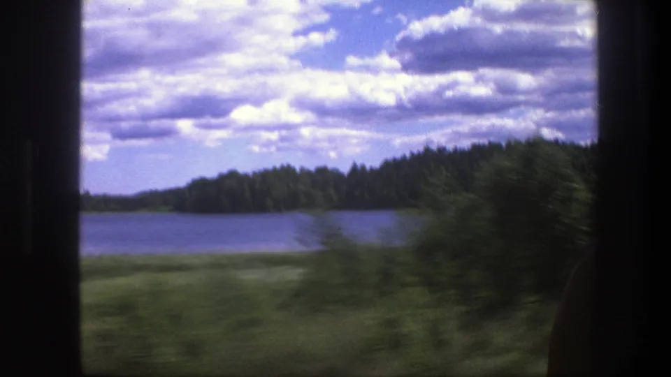 1975 Sweden home-movie frame showing a lake, wooded shoreline, cloudy sky, and motion-blurred greenery through a vehicle window.