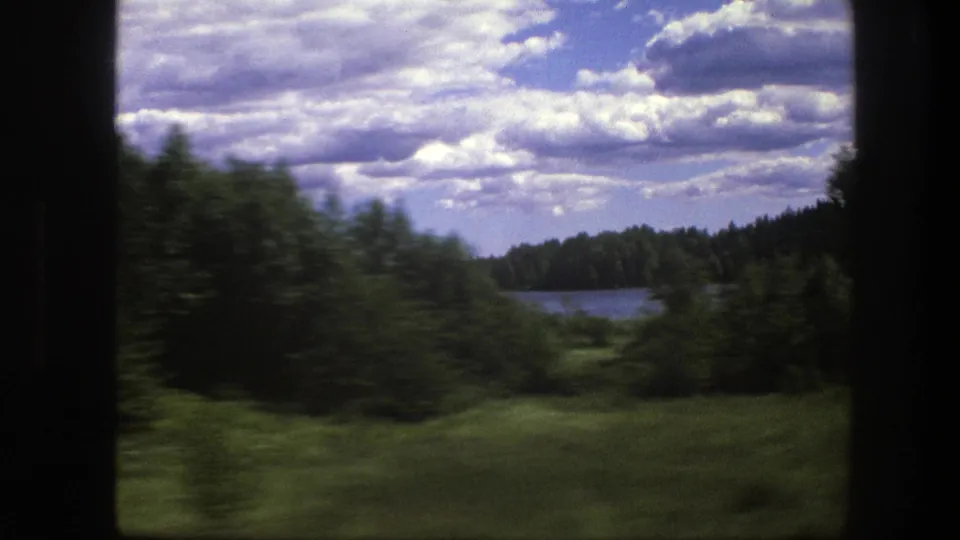 Window-framed 8mm view of rural Sweden with green field, bushes, a lake edge, distant forest, and layered clouds.