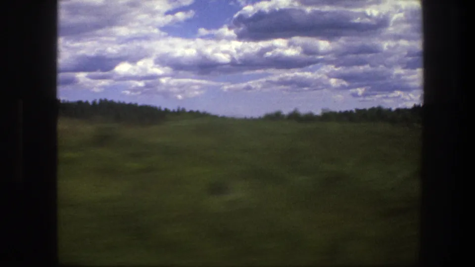 1975 Swedish rural landscape seen from a moving train, with open grassland, a low forest line, and scattered clouds across the sky.