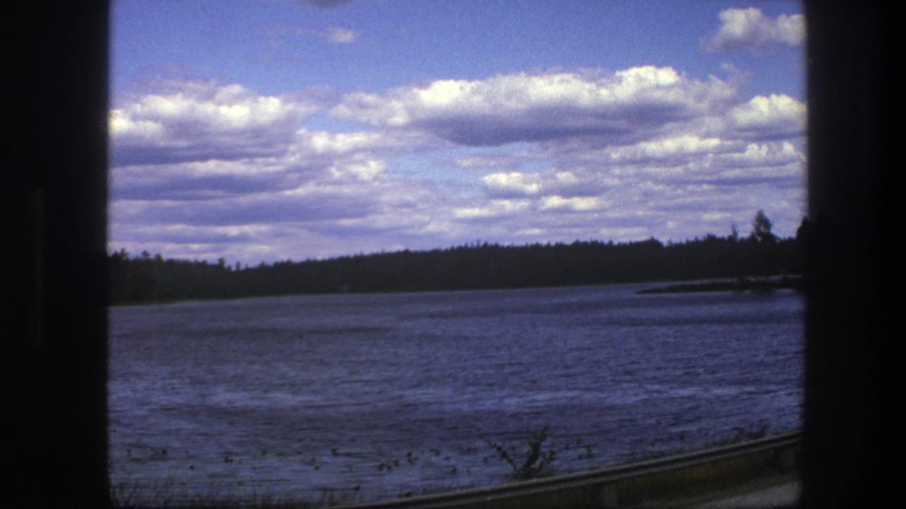 Train-window view across a lake toward a dark forest line under a cloud-filled blue sky.