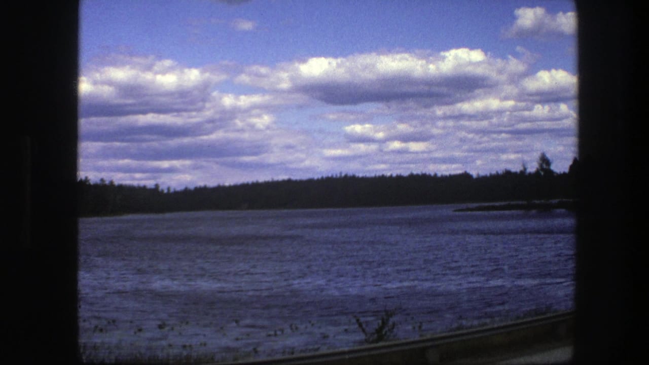 Lake and wooded shoreline seen through a train window, with broad clouds across the sky.