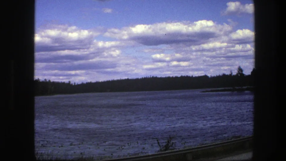 1975 8mm frame from Sweden showing a lake beside the rail line, distant trees, foreground reeds, and a partly cloudy sky.