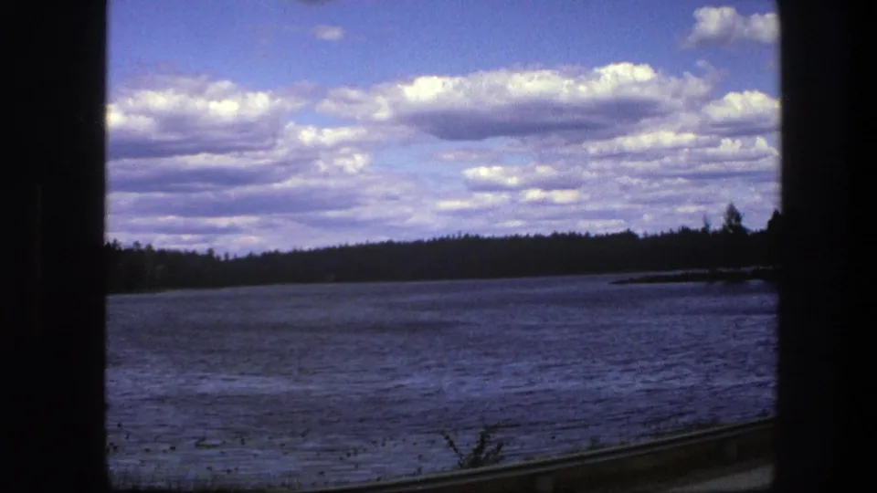 Swedish countryside in 1975 seen through a train window, with open lake water, low forest, and bright layered clouds.
