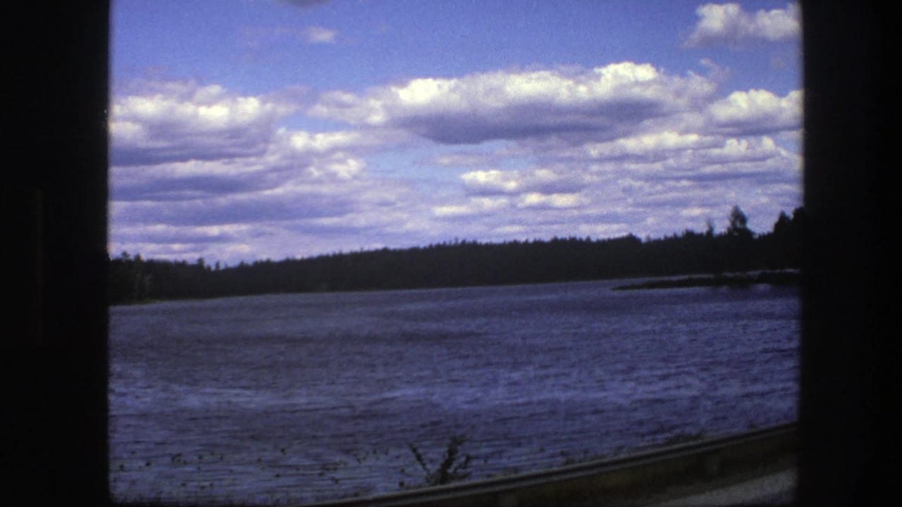 Blue lake seen past a vehicle window, with dark treeline and cloudy sky.