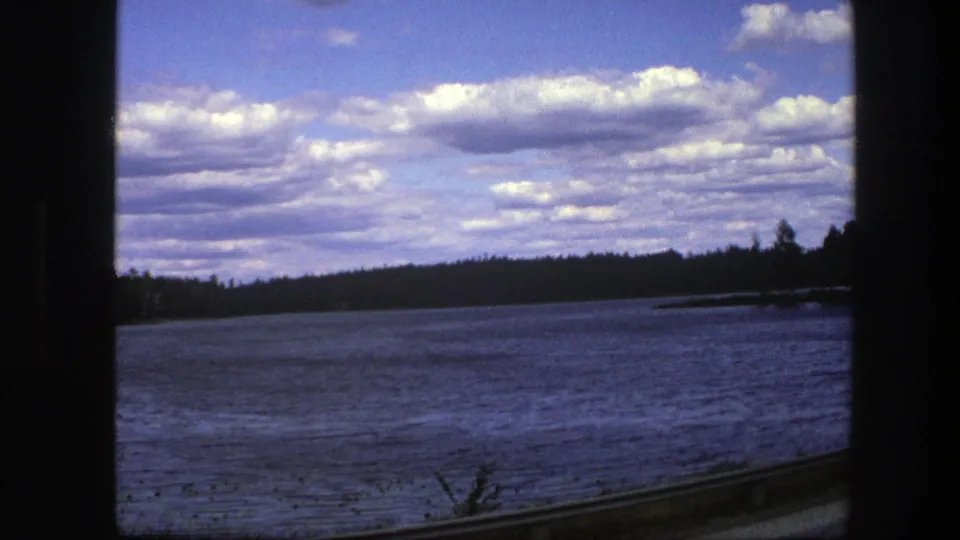 1975 Sweden 8mm frame showing a blue lake, distant evergreen shore, cloudy sky, and vehicle-window framing.