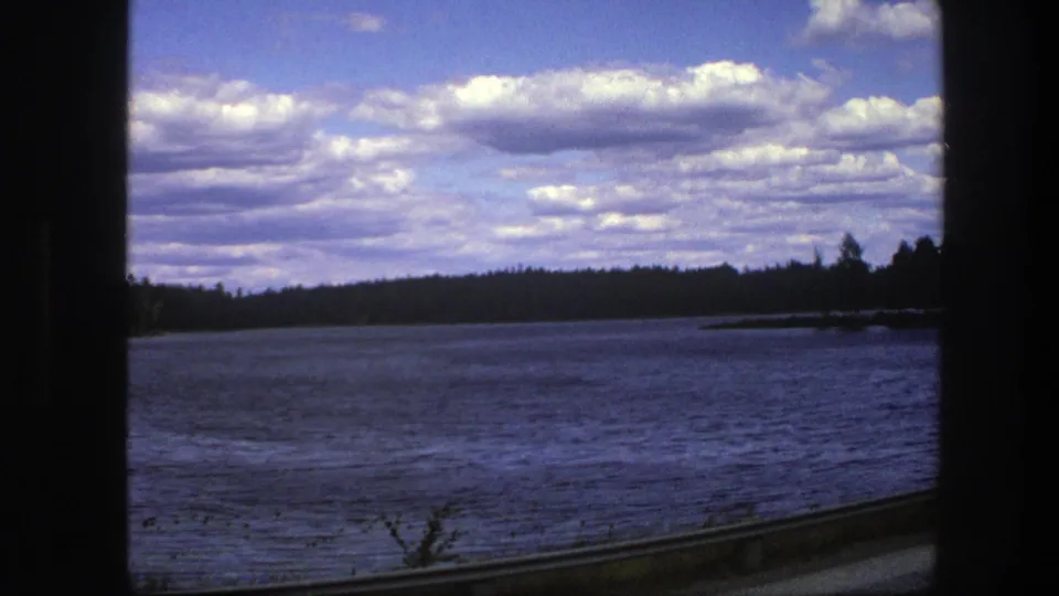 1975 Sweden home-movie frame of a wide lake, evergreen shoreline, cloud-filled sky, and car-window border.