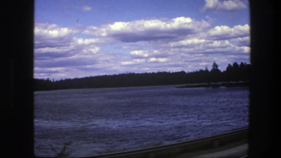 1975 Sweden 8mm frame showing open water, dark tree line, clouds, and the edge of a roadside vehicle view.