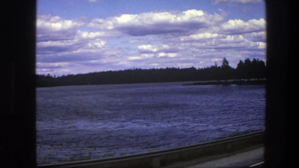 1975 Sweden travel frame with a broad blue lake, evergreen shore, scattered clouds, and dark car-window framing.