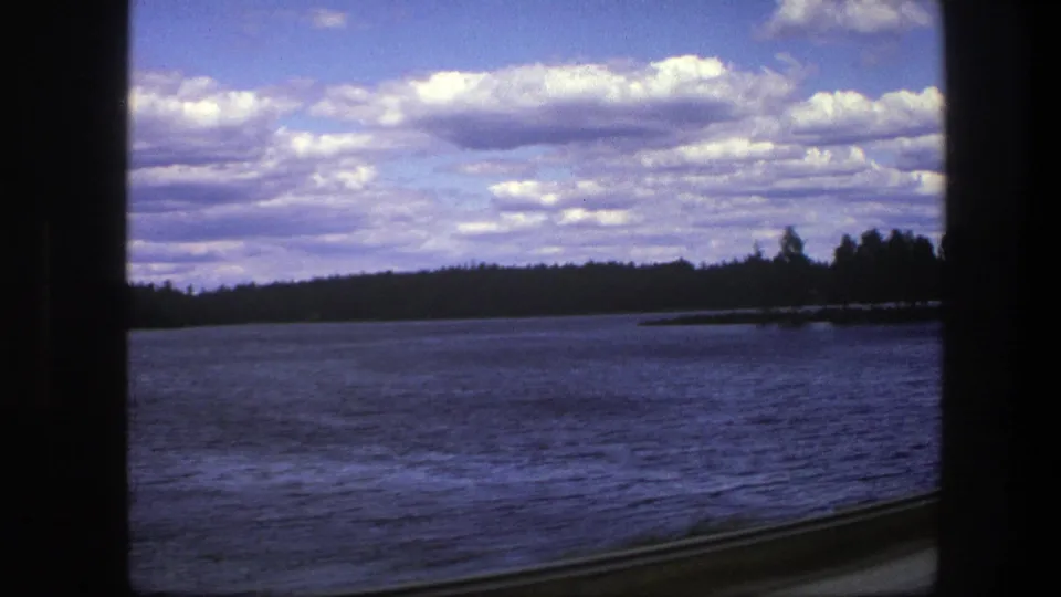 1975 Sweden 8mm frame showing a blue lake, wooded shoreline, cloudy sky, dark window borders, and a roadside guardrail from a moving vehicle.