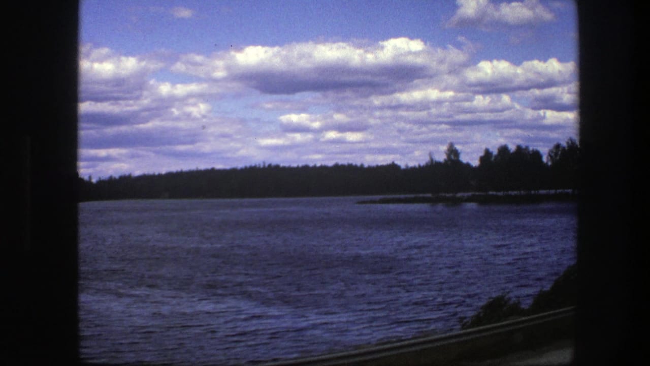 1975 Sweden 8mm frame of lake water and a low wooded shoreline seen past a dark window frame, with clouds overhead and guardrail below.