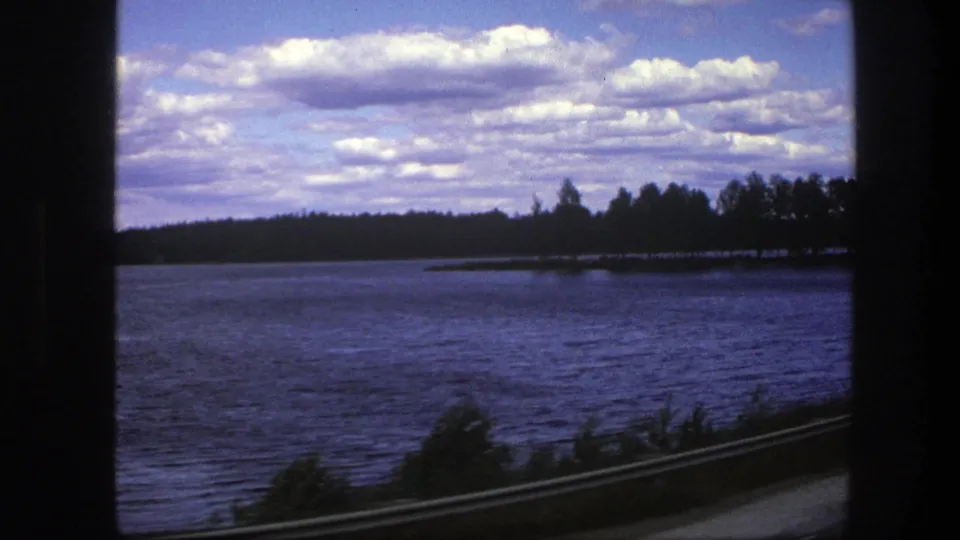 1975 Sweden 8mm frame showing a broad lake, dark window border, distant trees, cloudy sky, guardrail, and blurred roadside vegetation.