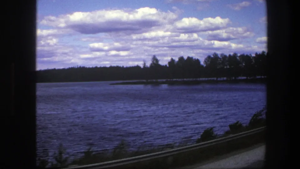 1975 Sweden 8mm frame with blue lake water, wooded shoreline, scattered clouds, a dark window edge, guardrail, and roadside plants in motion.
